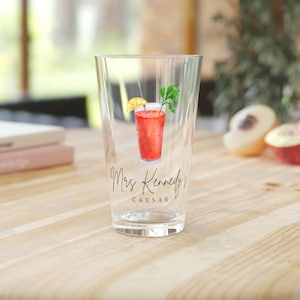 May include: A clear glass featuring a Caesar cocktail illustration. The drink is red with a lemon slice and celery garnish. The text "Mrs. Kennedy's Caesar" is written in cursive below the drink. The glass is on a wooden surface.