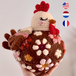 Może przedstawiać: Ręcznie robiona szydełkowa kura z brązowym korpusem ozdobionym kwiatowymi wzorami w odcieniach różu, bieli i pomarańczy. Kura ma kremową głowę, czerwony grzebień i korale oraz zamknięte oczy, tworząc kapryśny i uroczy design.