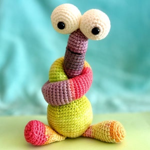 Könnte beinhalten: Ein farbenfrohes Häkelspielzeug mit großen weißen Augen und einem langen, gekräuselten Körper. Das Spielzeug ist aus verschiedenen Farbtönen von Rosa, Grün und Lila gefertigt.