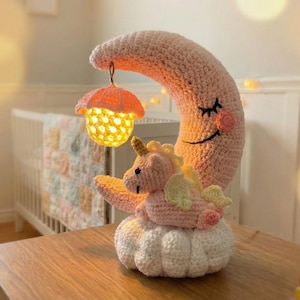 Peut inclure: Lampe de chambre d'enfant au crochet représentant un croissant de lune rose avec un visage souriant, une licorne et une base en forme de nuage. Une petite lampe orange en forme de champignon est suspendue à la lune. La lampe est sur une table en bois.