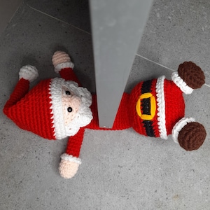 Peut inclure: Un protège-porte en crochet représentant le Père Noël en rouge, blanc et noir. Le protège-porte est posé sur un sol en carrelage gris avec un chambranle de porte gris en arrière-plan.