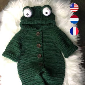 May include: A dark green, crocheted baby jumpsuit with a frog design. The hooded jumpsuit features large white and black eyes, a button-up front, and a ribbed texture. The outfit is designed to resemble a frog.