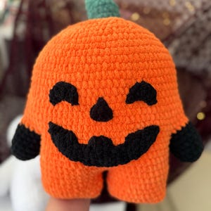 May include: A plush orange pumpkin toy made of soft yarn with a green stem. The pumpkin features a black crocheted face with two closed eyes, a small triangular nose, and a wide, jagged, smiling mouth. It has two black arms.