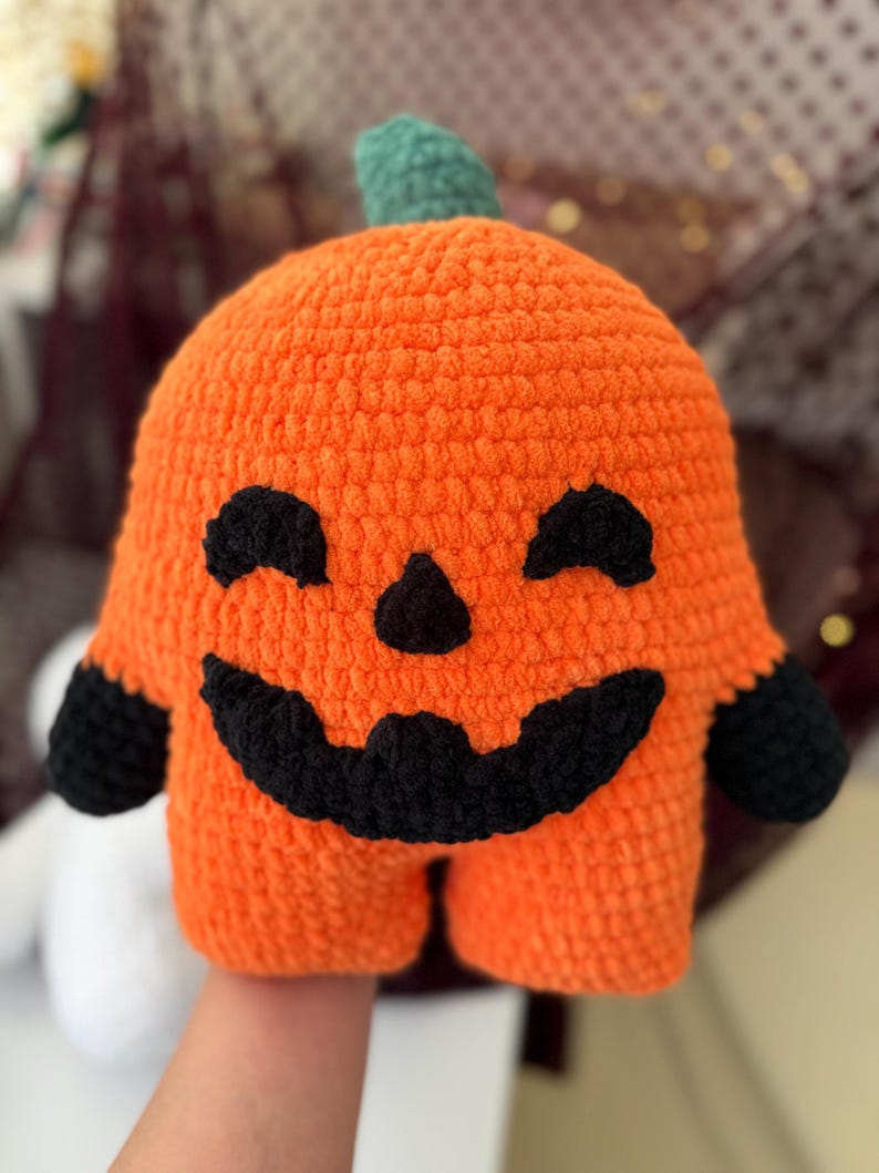 May include: A plush orange pumpkin toy made of soft yarn with a green stem. The pumpkin features a black crocheted face with two closed eyes, a small triangular nose, and a wide, jagged, smiling mouth. It has two black arms.