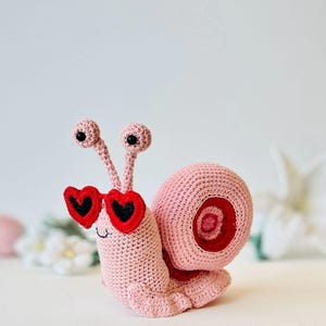 Peut inclure: Un escargot en crochet rose portant des lunettes de soleil en forme de c&oelig;ur rouge. L'escargot a des yeux noirs et une coquille rose avec un tourbillon rouge.