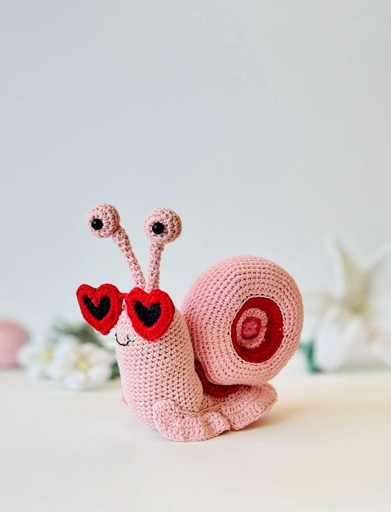 Peut inclure: Un escargot en crochet rose portant des lunettes de soleil en forme de c&oelig;ur rouge. L'escargot a des yeux noirs et une coquille rose avec un tourbillon rouge.