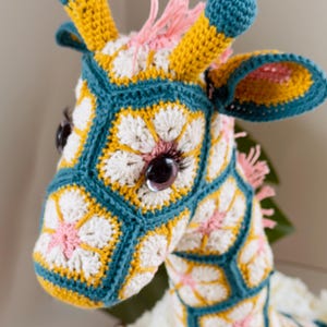 May include: A crochet giraffe stuffed animal constructed from hexagonal floral motifs in teal, yellow, white, and pink. The giraffe features dark brown plastic eyes with long eyelashes, two teal-tipped horns, and a pink yarn mane along the neck.