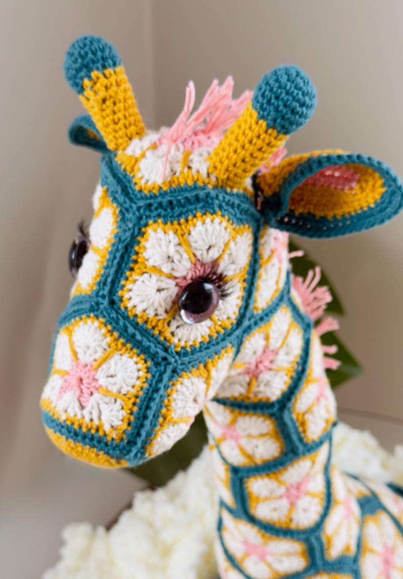 May include: A crochet giraffe stuffed animal constructed from hexagonal floral motifs in teal, yellow, white, and pink. The giraffe features dark brown plastic eyes with long eyelashes, two teal-tipped horns, and a pink yarn mane along the neck.