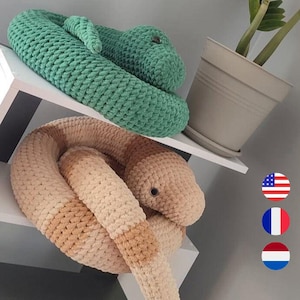Könnte beinhalten: Zwei Plüsch-Schlangenspielzeuge. Eines ist grün, das andere ist beige und braun. Die Schlangen bestehen aus einem weichen, strukturierten Material und sind auf einem weißen Regal aufgerollt. Die Schlangen haben schwarze Knopfaugen.