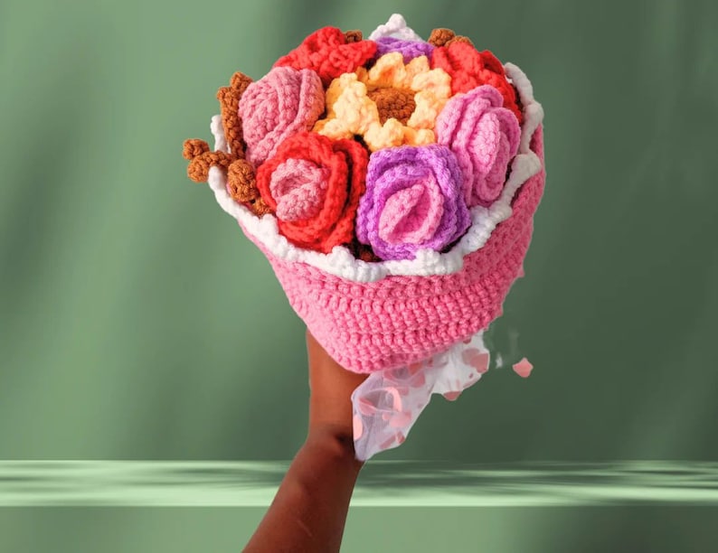 May include: A crocheted bouquet of flowers in shades of pink, red, purple, and yellow. The bouquet is wrapped in a pink crocheted paper with white trim.