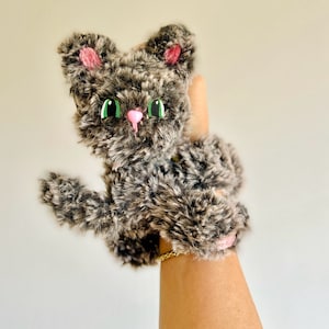 Può includere: Un giocattolo di peluche grigio a forma di gatto con orecchie rosa e occhi verdi. Il gatto è tenuto da una mano che indossa un braccialetto dorato.