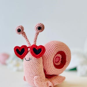 Peut inclure: Un escargot en crochet rose portant des lunettes de soleil en forme de c&oelig;ur rouge. L'escargot a des yeux noirs et une bouche souriante.