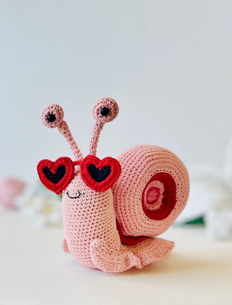 Peut inclure: Un escargot en crochet rose portant des lunettes de soleil en forme de c&oelig;ur rouge. L'escargot a des yeux noirs et une bouche souriante.