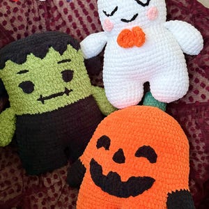 May include: Three plush crochet toys featuring a green monster with black hair, a white ghost with pink cheeks and an orange bow, and an orange pumpkin with a black face. Each figure is crafted from soft yarn with embroidered facial details.