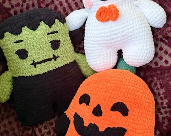 May include: Three plush crochet toys featuring a green monster with black hair, a white ghost with pink cheeks and an orange bow, and an orange pumpkin with a black face. Each figure is crafted from soft yarn with embroidered facial details.