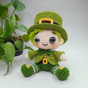 以下が含まれることがあります： 手作りのかぎ針編みのレプラコーン人形。人形は、黄色のバックルが付いた緑色の帽子、緑色のジャケット、白いアクセントが付いた緑色のズボンを着用しています。人形は緑色の髪、バラ色の頬、大きくて表現力豊かな目をしています。人形は座っています。