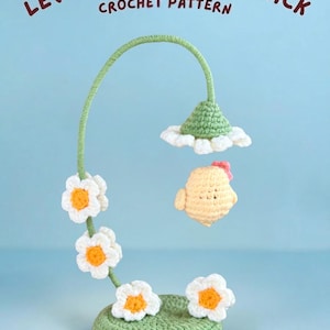 Op de afbeelding: Een gehaakte lentedecoratie met een kuiken en bloemen. Het ontwerp omvat een groene basis, een gebogen stengel met witte en gele bloemen en een zwevend kuiken. De tekst "LEVITATING SPRING CHICK CROCHET PATTERN" staat er boven.
