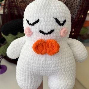 May include: A white crocheted plush toy with a rounded head and body. It features embroidered black closed eyes and a small smile, pink circular cheeks, and a bright orange bow tie made of two yarn circles centered on the chest.