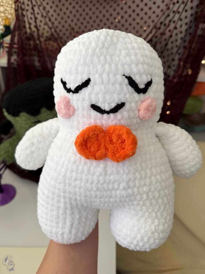May include: A white crocheted plush toy with a rounded head and body. It features embroidered black closed eyes and a small smile, pink circular cheeks, and a bright orange bow tie made of two yarn circles centered on the chest.