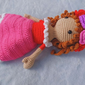May include: A crocheted doll with brown hair, wearing a pink dress with white trim and a red bodice. The doll is wearing a flower crown with pink, purple, and red flowers.