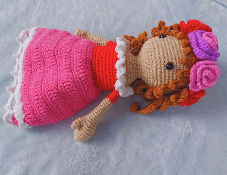 May include: A crocheted doll with brown hair, wearing a pink dress with white trim and a red bodice. The doll is wearing a flower crown with pink, purple, and red flowers.