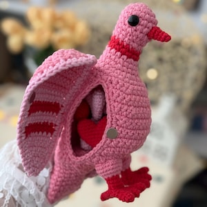 May include: A handmade pink crochet pigeon with red accents. The pigeon has open wings and a red heart inside. The bird's beak and feet are red. The pigeon is made of soft yarn and has a button closure.