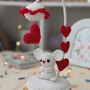 Peut inclure: Mobile crochet fait main avec une figurine de souris blanche, de grands cœurs rouges et un dessus en forme de lampe blanche avec une bordure rouge. Le mobile est posé sur une base en bois.