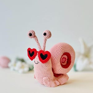 Peut inclure: Un escargot en crochet rose portant des lunettes de soleil en forme de c&oelig;ur rouge. L'escargot a des yeux noirs et un petit sourire.