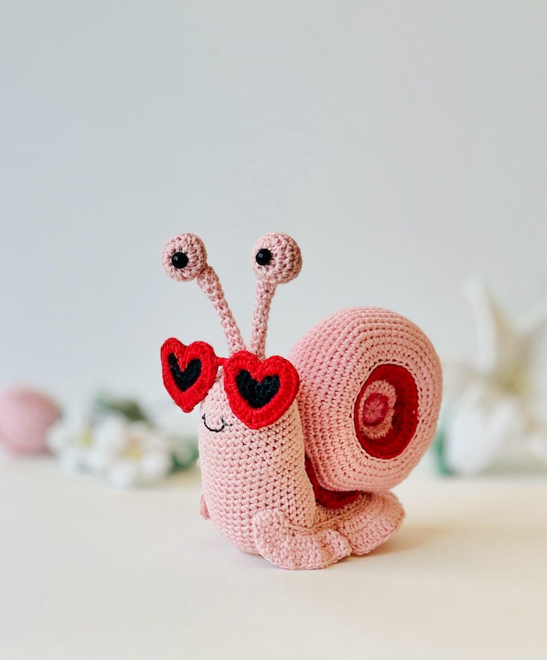 Peut inclure: Un escargot en crochet rose portant des lunettes de soleil en forme de c&oelig;ur rouge. L'escargot a des yeux noirs et un petit sourire.