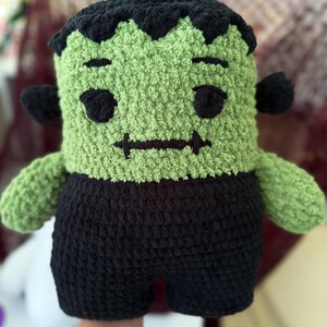 May include: A plush crochet doll featuring a green head with black hair, black eyes, and a stitched mouth. The doll has black bolts on the sides of its head and wears black pants. The texture is soft and fuzzy, crafted from thick yarn.