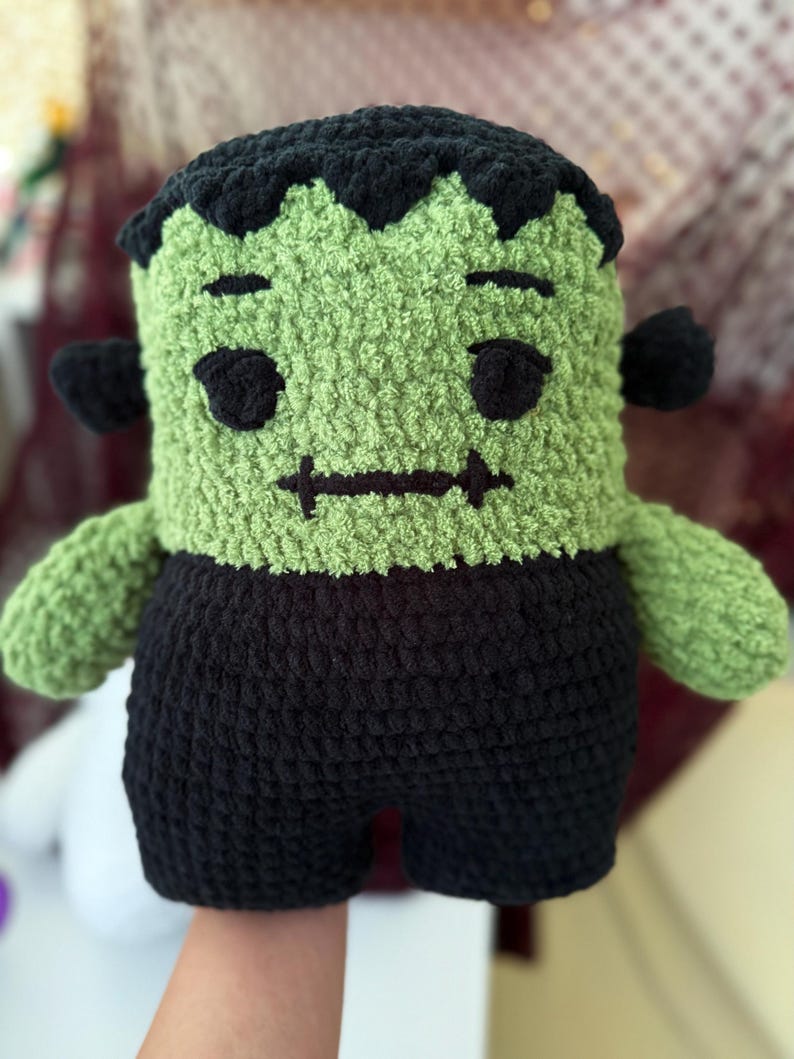May include: A plush crochet doll featuring a green head with black hair, black eyes, and a stitched mouth. The doll has black bolts on the sides of its head and wears black pants. The texture is soft and fuzzy, crafted from thick yarn.