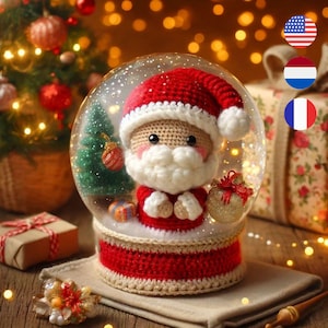 Puede incluir: Una figurita de Papá Noel de crochet dentro de una bola de nieve de vidrio con una base de crochet roja y blanca. La bola de nieve está decorada con un pequeño árbol de Navidad verde y un regalo dorado.