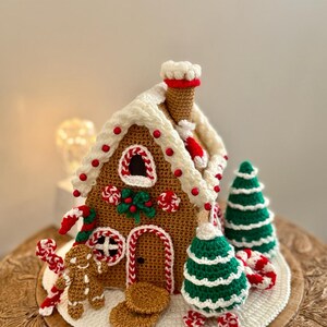 May include: A crocheted gingerbread house with white icing, red candies, and a chimney. The house is decorated with a gingerbread man, candy canes, and green Christmas trees. The house sits on a white crocheted base.