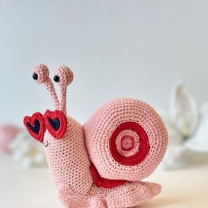 Peut inclure: Un escargot en crochet rose portant des lunettes de soleil en forme de c&oelig;ur rouge. L'escargot a une coquille rose avec une bande rouge et blanche.
