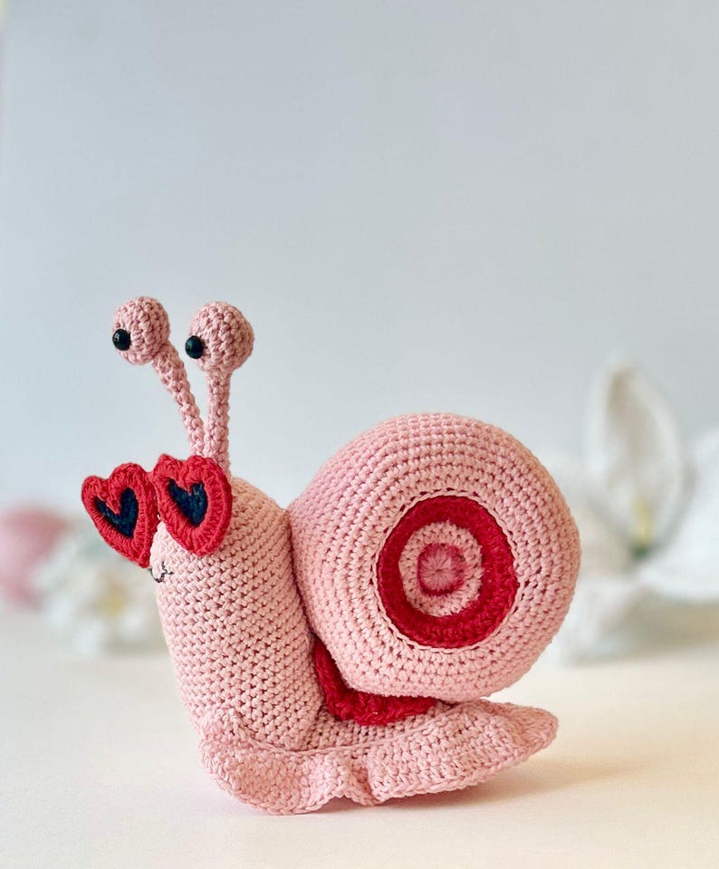Peut inclure: Un escargot en crochet rose portant des lunettes de soleil en forme de c&oelig;ur rouge. L'escargot a une coquille rose avec une bande rouge et blanche.