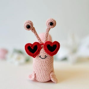 Peut inclure: Un escargot en crochet rose portant des lunettes de soleil en forme de c&oelig;ur rouge. L'escargot a des yeux noirs et un petit sourire.