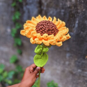 May include: A handmade crochet sunflower with yellow petals and a brown center. The stem is green with green leaves. The flower is held in a person's hand against a blurred background.