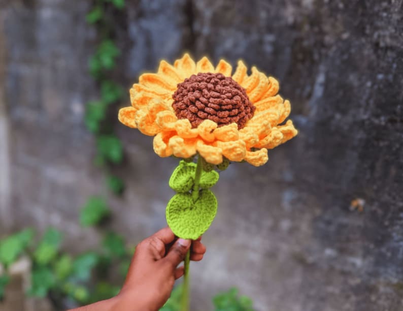 May include: A handmade crochet sunflower with yellow petals and a brown center. The stem is green with green leaves. The flower is held in a person's hand against a blurred background.
