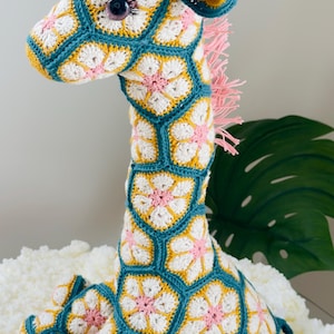 May include: A handcrafted crochet giraffe toy constructed from interconnected hexagonal motifs. The pattern features white and pink floral centers outlined in teal yarn, with yellow accents on the horns and ears. It has a pink yarn mane and tail.