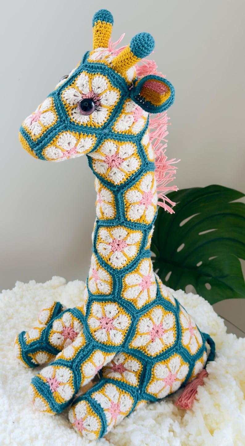 May include: A handcrafted crochet giraffe toy constructed from interconnected hexagonal motifs. The pattern features white and pink floral centers outlined in teal yarn, with yellow accents on the horns and ears. It has a pink yarn mane and tail.