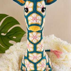 May include: A crochet giraffe stuffed animal constructed from hexagonal floral motifs in teal, white, yellow, and pink yarn. The giraffe features two small horns, large black plastic eyes with eyelashes, and a pink yarn tuft on its head and tail.