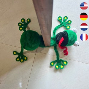 Peut inclure: Un arrêt de porte en forme de grenouille crocheté de couleur verte avec des accents jaunes et une langue rouge. La grenouille est positionnée pour maintenir une porte ouverte.