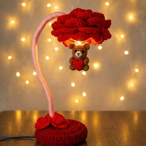 Puede incluir: Una lámpara de ganchillo hecha a mano con una pantalla y base en forma de rosa roja. La lámpara presenta un tallo rosa y un pequeño oso de peluche con un corazón. El fondo tiene luces suaves y brillantes.