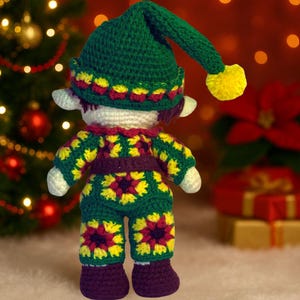 May include: A handmade crochet elf doll. It is wearing a green hat with a yellow bobble, and a matching outfit with yellow and red floral patterns. The doll has white ears and is in front of a Christmas tree.