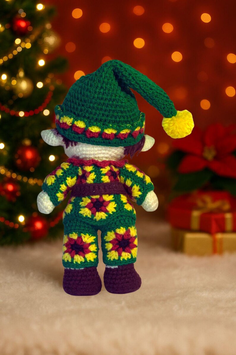 May include: A handmade crochet elf doll. It is wearing a green hat with a yellow bobble, and a matching outfit with yellow and red floral patterns. The doll has white ears and is in front of a Christmas tree.