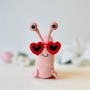 Peut inclure: Un escargot en crochet rose portant des lunettes de soleil en forme de c&oelig;ur rouge. L'escargot a des yeux noirs et un petit sourire.