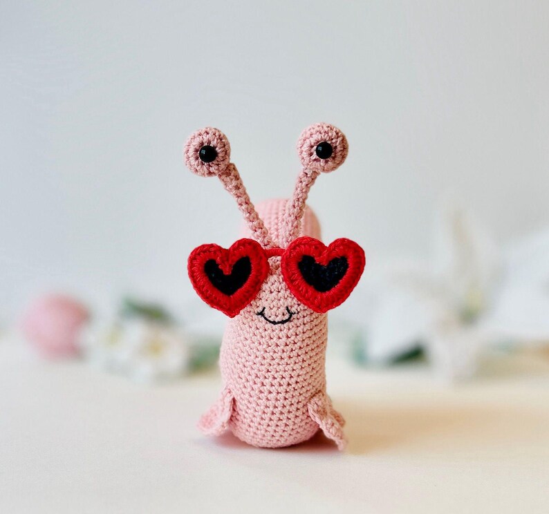 Peut inclure: Un escargot en crochet rose portant des lunettes de soleil en forme de c&oelig;ur rouge. L'escargot a des yeux noirs et un petit sourire.