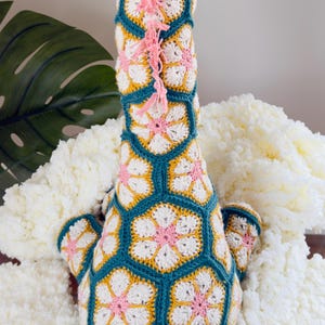 May include: A crochet giraffe toy constructed from hexagonal floral motifs in teal, white, yellow, and pink yarn. The giraffe features a long neck, two small horns, and a braided tail with a pink tassel. A pink yarn mane runs down the neck.