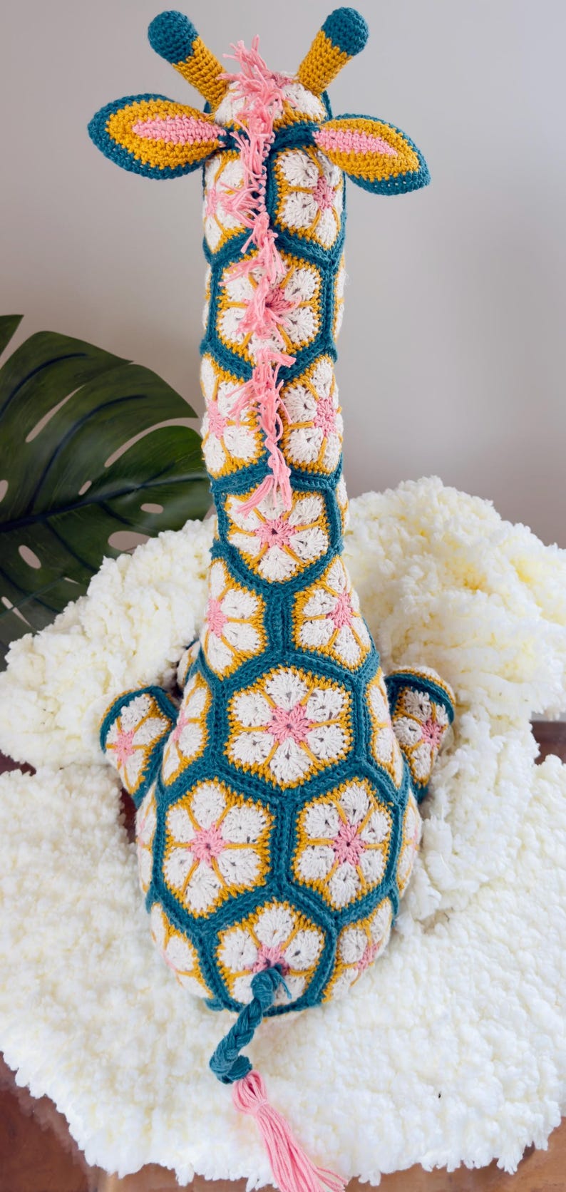 May include: A crochet giraffe toy constructed from hexagonal floral motifs in teal, white, yellow, and pink yarn. The giraffe features a long neck, two small horns, and a braided tail with a pink tassel. A pink yarn mane runs down the neck.