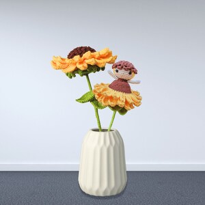 May include: A cream-colored vase holds two crocheted sunflowers with green stems and leaves. A small crocheted doll with a brown dress and a pink head is perched on one of the flowers. The sunflowers are yellow and brown.